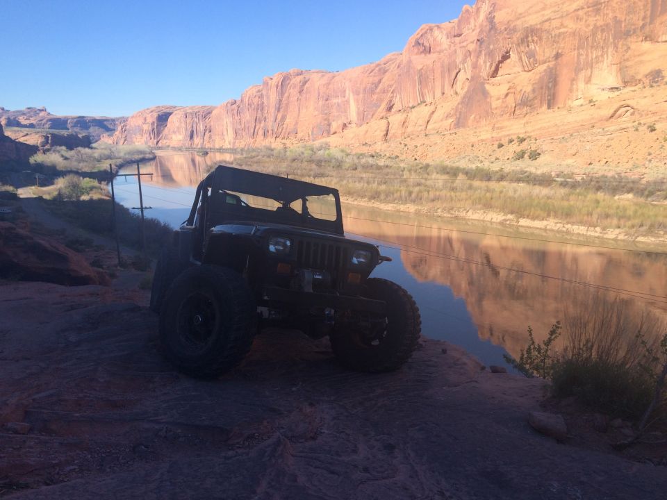 Dark Jeep YJ parked on rocks by a river with red cliffs.