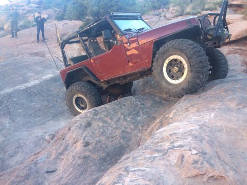 1995 Jeep Wrangler - A red Jeep Wrangler is navigating a steep, rocky incline off-road. The vehicle has large, aggressive tires and a roll cage, indicating it i…
