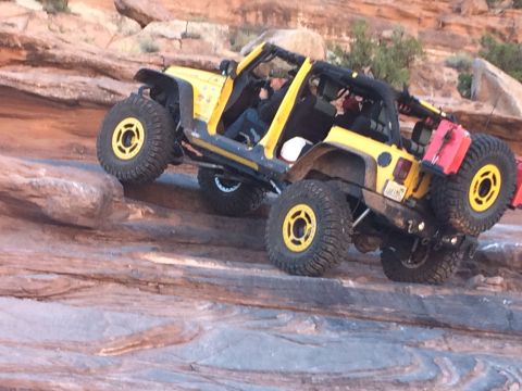 1995 Jeep Wrangler - A bright yellow Jeep Wrangler is shown off-roading on a rocky, desert terrain. The vehicle is tilted at an extreme angle as it navigates ov…