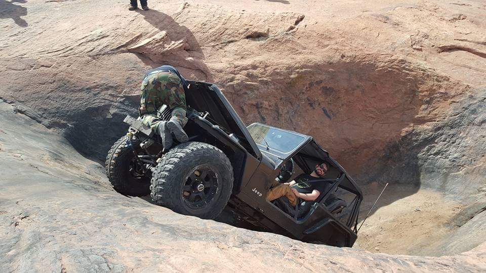 Black Jeep YJ tilted on rocky terrain, one person on the exterior, one in the driver's seat.