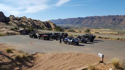 1995 Jeep Wrangler - A group of off-road vehicles, including several Jeeps and UTVs, are parked in a lot with a scenic desert landscape and mountains in the bac…