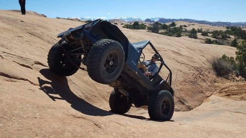 1995 Jeep Wrangler - A dark-colored Jeep YJ is shown at a steep angle, climbing a rocky, desert terrain. The driver, wearing sunglasses and a plaid shirt, is vi…