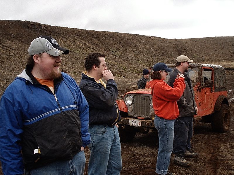 Muddy orange open-top off-road Jeep with European plate in barren hills, four people standing nearby