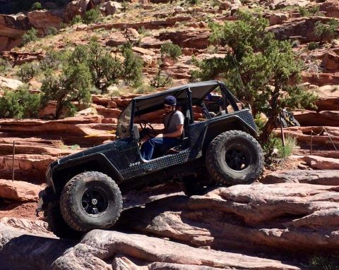1995 Jeep Wrangler - A black Jeep YJ is navigating a rocky, off-road trail with steep red rock formations and sparse green trees in the background. The driver i…