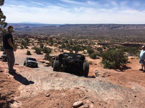 1995 Jeep Wrangler - Two modified Jeep Wranglers navigate a rocky, off-road trail with a vast desert canyon in the background. A person stands on the left, obse…