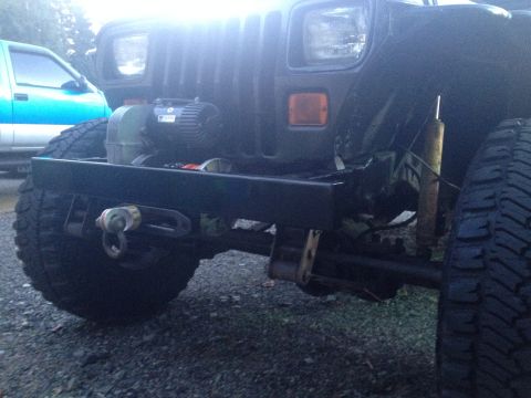 1995 Jeep Wrangler - A close-up, low-angle shot of the front bumper and winch of a dark-colored Jeep YJ. A blue pickup truck is visible in the background.