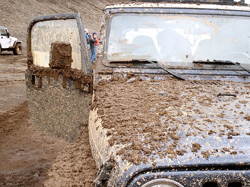 Mud-covered dark SUV front quarter at an off-road muddy site with white Jeep behind
