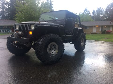 1995 Jeep Wrangler - A black Jeep YJ sits parked on a wet asphalt driveway in front of a suburban house. The vehicle features large off-road tires, a winch moun…