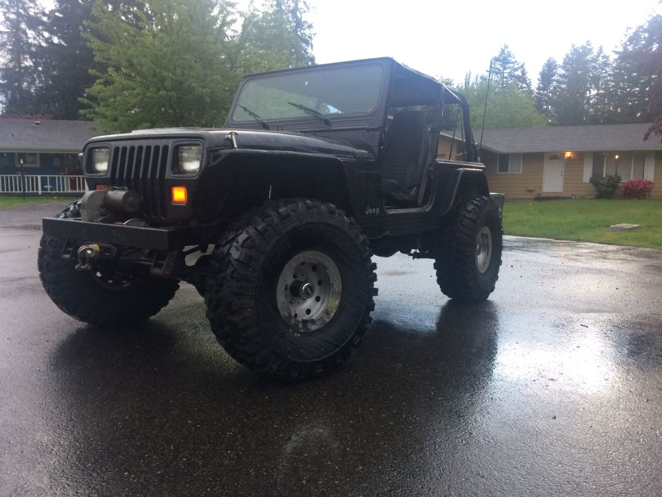 Black Jeep YJ with large tires and winch on a wet driveway.