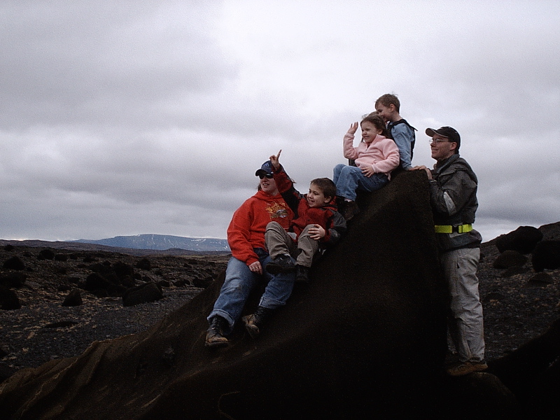 Family of five sitting on a large dark rock in a rocky landscape under gray clouds.