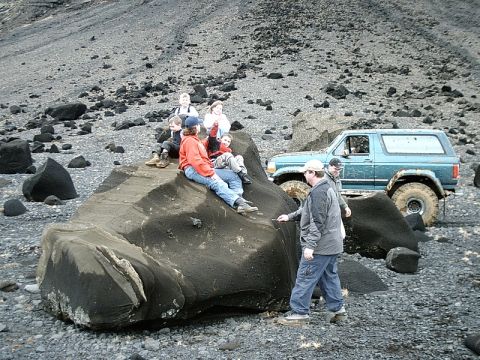 1996 Ford Bronco - A group of people take a break on a large rock formation, with a blue SUV nearby in a barren, rocky landscape.