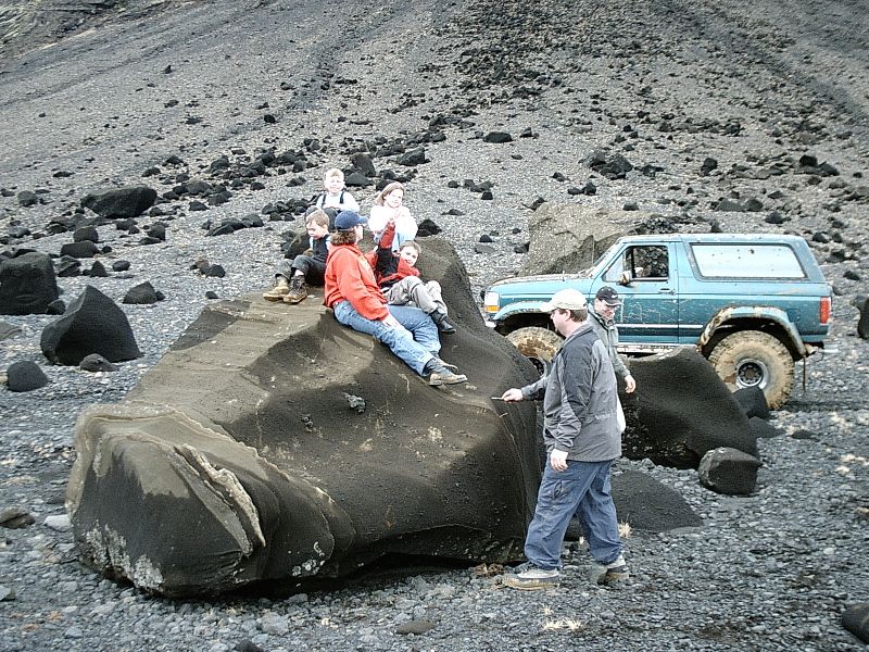 Teal Ford Bronco SUV parked on black volcanic gravel with people on a large rock nearby