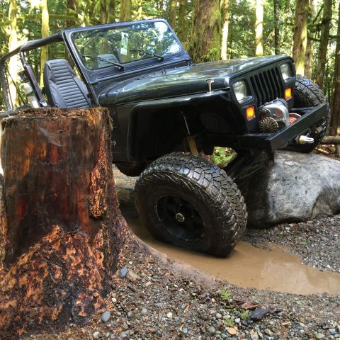 1995 Jeep Wrangler - A black Jeep YJ is navigating a muddy trail in a forest, with its front tire resting on a large rock and its rear tire in a puddle next to…