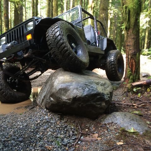 1995 Jeep Wrangler - A black Jeep YJ is shown from a low angle, navigating over a large rock on a wooded trail. The vehicle's large, knobby tires are visible, a…