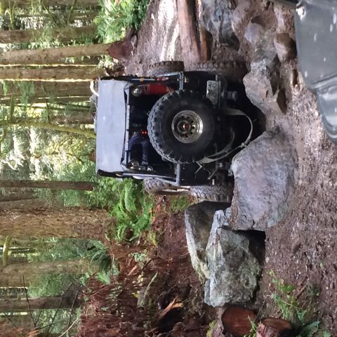 1995 Jeep Wrangler - A lifted Jeep YJ navigates a rocky, wooded trail. The vehicle is shown from a rear-side angle, with its spare tire prominently displayed on…