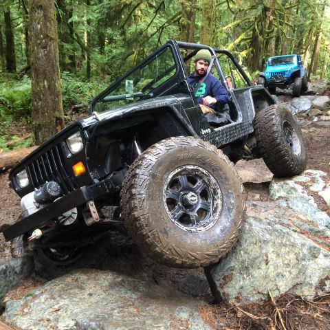 1995 Jeep Wrangler - A black Jeep YJ is navigating a rocky, wooded trail, with a blue Jeep visible in the background. The driver, wearing a green beanie and a b…
