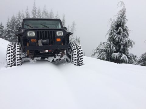 1995 Jeep Wrangler - A black 1995 Jeep YJ navigates a snowy mountain pass, its tires kicking up snow as it moves through the winter landscape.