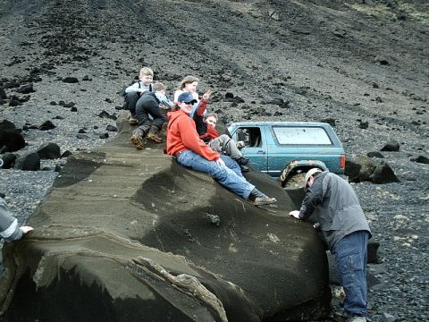 1996 Ford Bronco - A blue 1996 Ford Bronco is parked on a rocky, barren landscape while a group of people sit on a large rock nearby.
