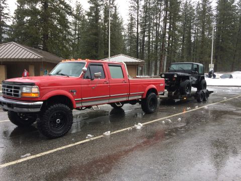 1995 Jeep Wrangler - A red Ford F-Series truck tows a black Jeep Wrangler on a trailer in a snowy, wet parking lot.