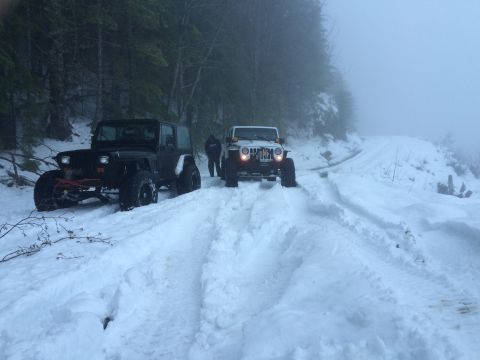 1995 Jeep Wrangler - Two Jeep Wranglers navigate a snow-covered mountain road, with the black YJ in the foreground and a white JK behind it.