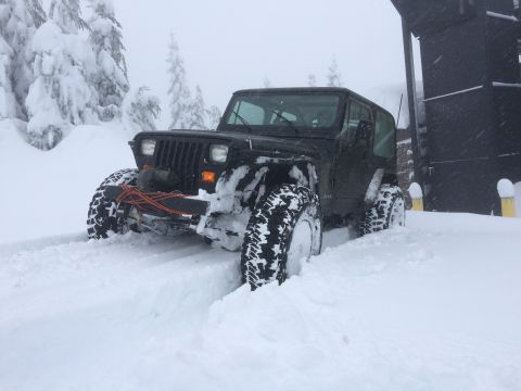 1995 Jeep Wrangler - A Jeep Wrangler navigates through a snowy landscape, with trees and a building visible in the background.