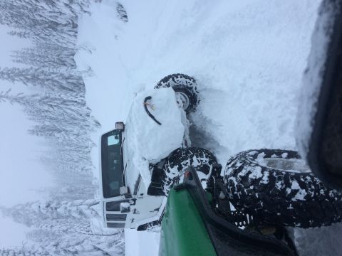 1995 Jeep Wrangler - A white utility vehicle with a snow plow is partially buried in snow, surrounded by snow-covered trees.