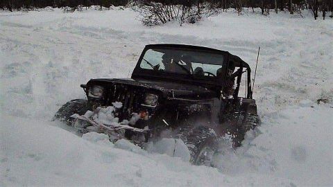 1995 Jeep Wrangler - A black Jeep Wrangler navigates through a snowy landscape, kicking up snow as it goes.