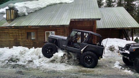 1995 Jeep Wrangler - A dark gray Jeep Wrangler YJ navigates a snow-covered area near a log cabin, with another Jeep visible in the background.