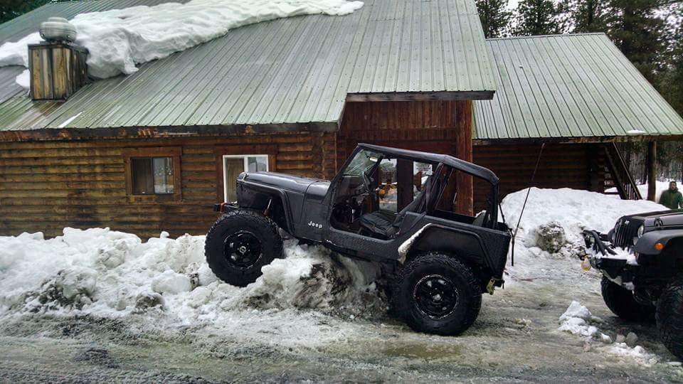 Dark gray Jeep Wrangler YJ with large tires stuck in snowdrift in front of log cabin