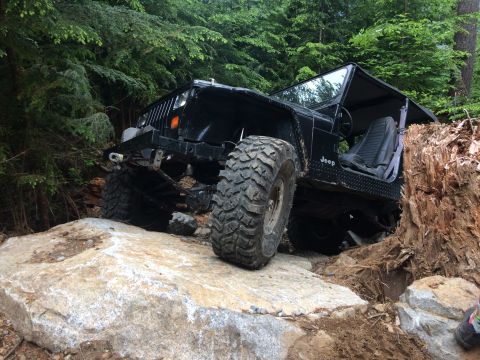 1995 Jeep Wrangler - A black Jeep Wrangler is shown off-roading on a rocky trail, with its tires positioned on large boulders.
