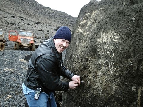 1996 Ford Bronco - A man carves graffiti into a large rock, with two off-road vehicles visible in the background on a desolate, rocky landscape.