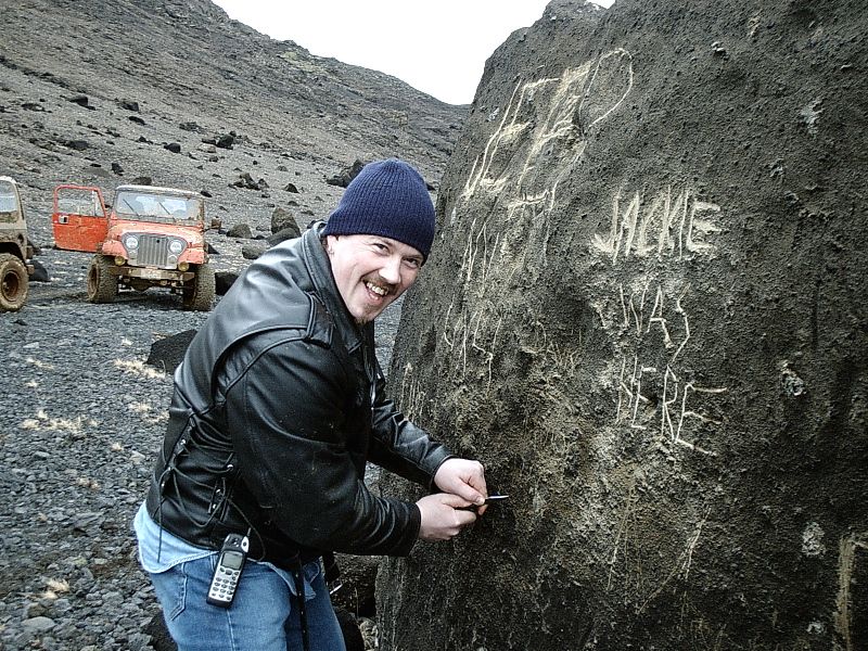 Man carving graffiti on a boulder with two rusty orange Jeeps parked on a rocky hillside
