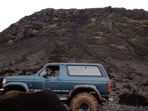 1996 Ford Bronco - A teal Ford Bronco is parked on a rocky, desolate landscape with a large, dark mountain dominating the background.