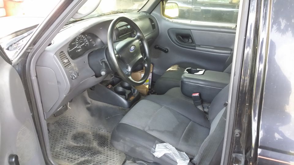 Interior of a black pickup viewed through open driver's door, gray dash, Ford steering wheel, manual shifter, cloth seats