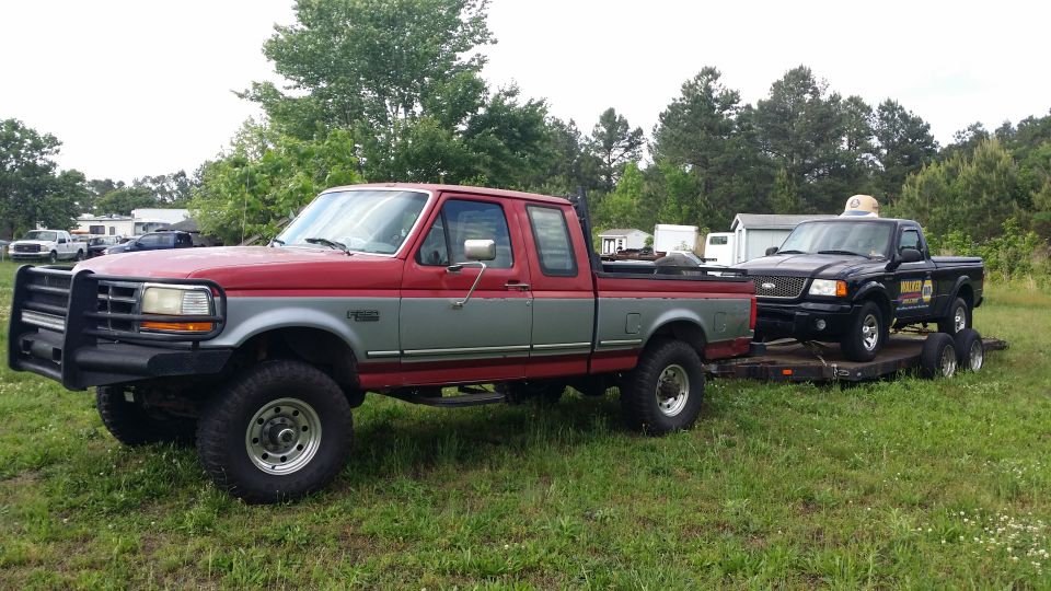 Red and silver pickup truck towing a black pickup truck on a trailer. Visible text on black truck