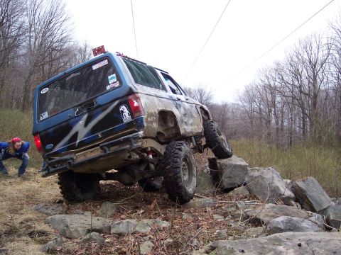 1978 Ford Bronco - A blue SUV navigates a rocky obstacle course on a wooded trail.