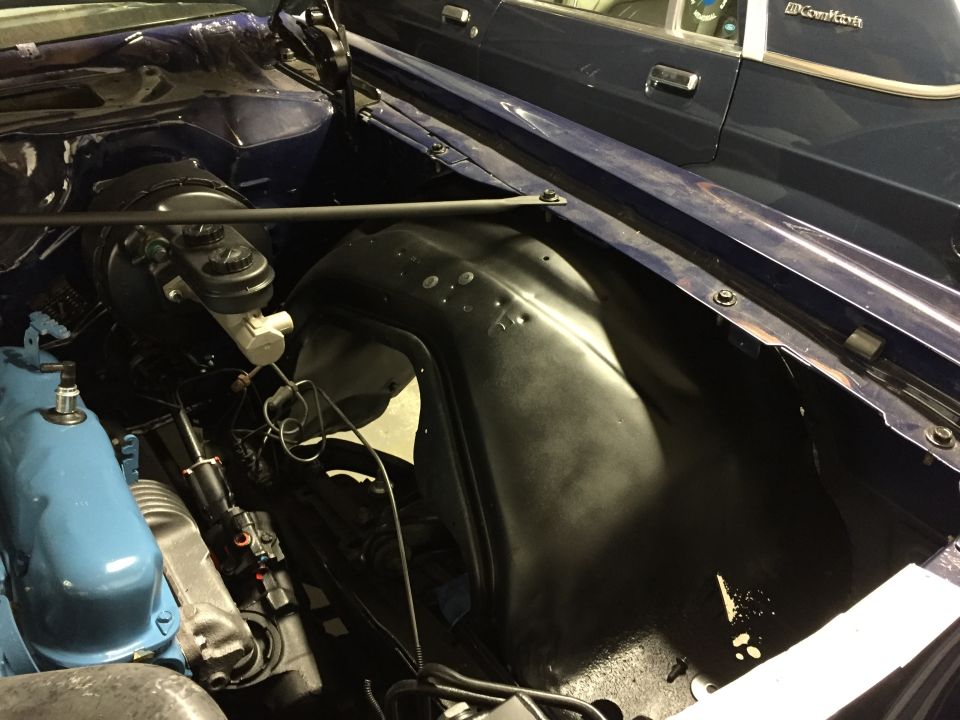Blue engine bay with visible brake master cylinder and inner fender well. Another dark blue car is visible in the background
