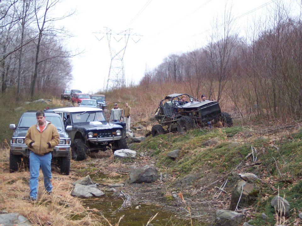 Several off-road vehicles, including a dark Bronco-like SUV and a roll cage buggy, navigate a wooded trail with a power line tower in the background