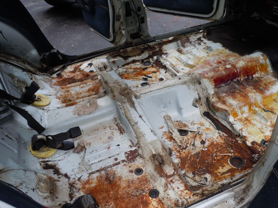 Car interior floor with extensive rust and damage, showing seatbelt anchors