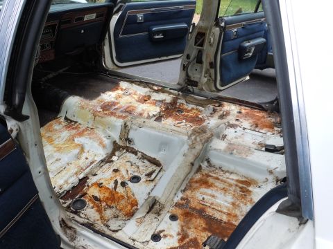 2001 Ford Crown Victoria - The interior of a car reveals extensive rust damage on the floor pans, with blue upholstery visible on the doors and dashboard.