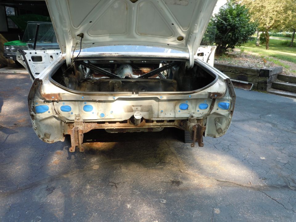 Car's trunk is open, showing the empty interior. Rear bumper area is stripped down, revealing rust and some blue tape