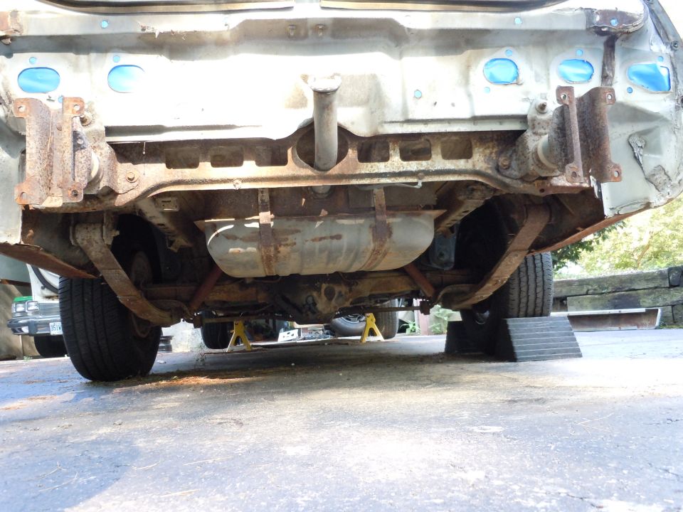 Rear undercarriage of a car showing fuel tank, leaf springs, and rusted frame components
