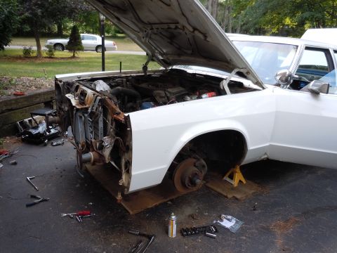 2001 Ford Crown Victoria - The front end of a white sedan is disassembled, with the hood open and engine components exposed. Tools are laid out on the driveway.