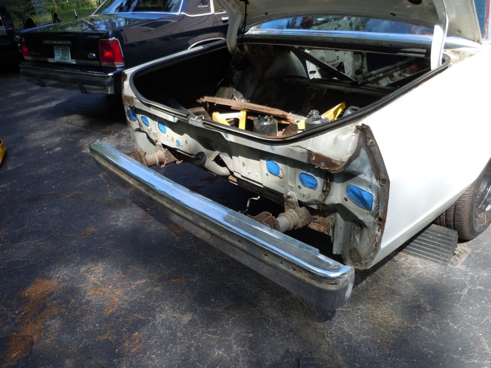 Rear bumper and damaged trunk area of a white car, with a black car in the background. Tools are visible in the trunk