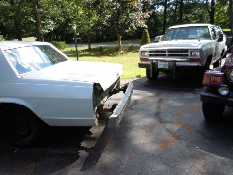 2001 Ford Crown Victoria - A white sedan with its front bumper missing sits on a driveway next to other vehicles.