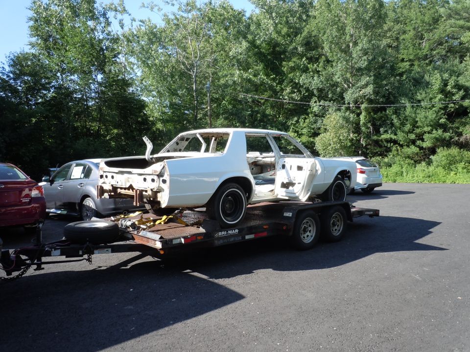 White sedan chassis on a trailer, doors open, stripped interior, black wheels, parked outdoors