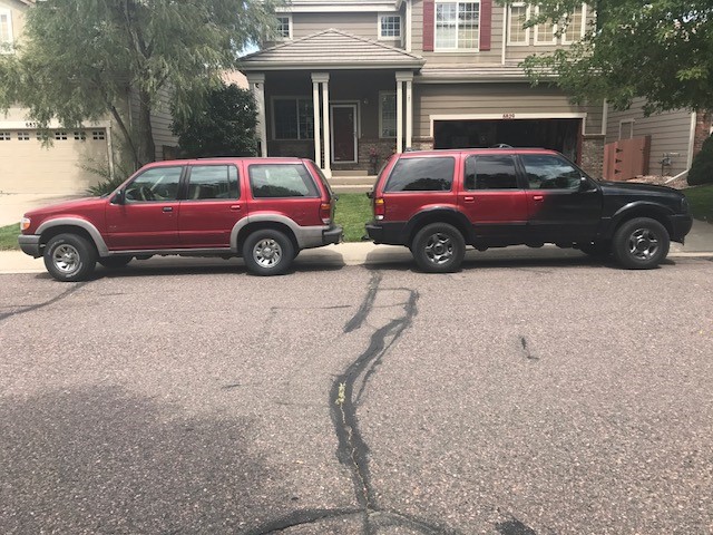 Two Ford Explorers parked side-by-side in front of a house. One is red, the other is red and black