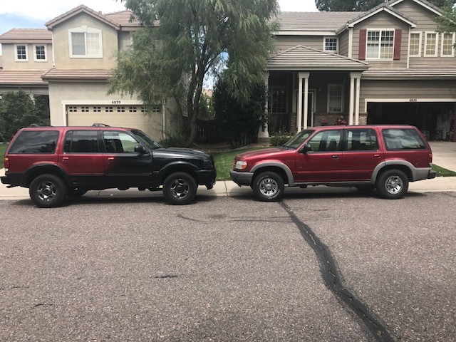 Two red Ford Explorers parked side-by-side on a street in front of houses. The one on the left has a black front end