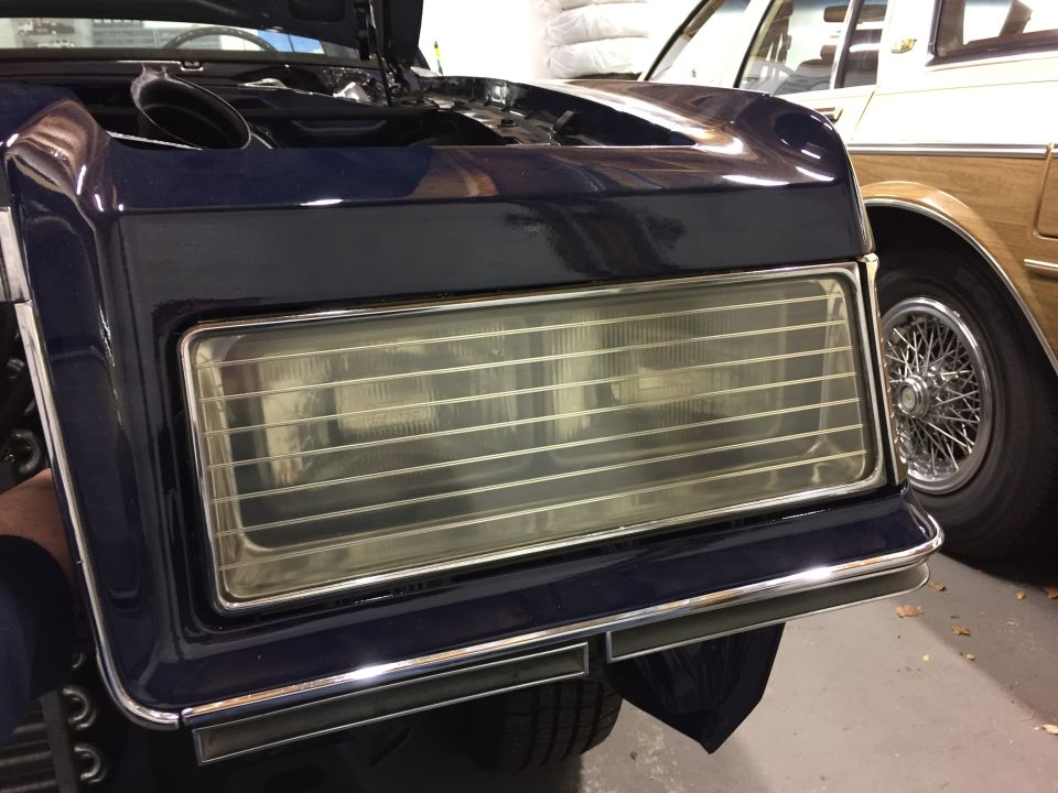 Dark blue car front with hood up, showing headlight assembly and chrome trim. Another car with wire wheels is to the right