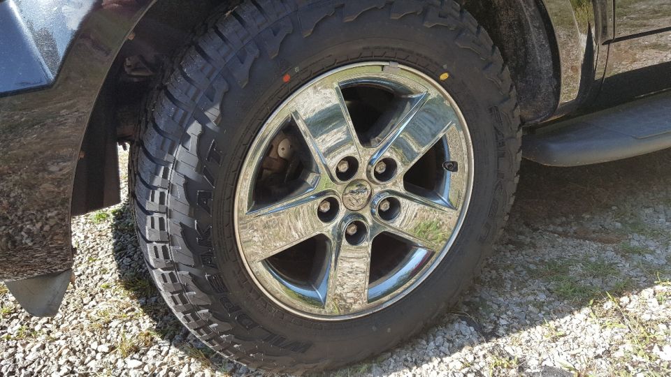 Chrome wheel with five spokes and a Ram logo, mounted on a tire. Part of a dark-colored vehicle is visible